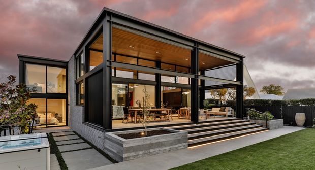 Contemporary two-storey residence viewed from the rear garden, featuring a strongly expressed black structural frame, extensive floor-to-ceiling glazing, warm timber-lined ceilings and walls, broad stepped decking, and an outdoor entertaining area. A lawn, paved garden path, raised concrete planters, fencing, and a partially visible pool or spa surround the house. The dusk sky adds a dramatic backdrop.