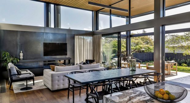 A sophisticated open-plan living and dining area with expansive floor-to-ceiling glazing opening toward a landscaped outdoor space. The room features a large light-grey sectional sofa, dark feature wall with a mounted television and built-in media joinery, a long dark dining table with black chairs, a marble-look kitchen island or benchtop in the foreground, and a warm timber slatted ceiling. Strong black structural framing and extensive glazing create a bright, contemporary indoor-outdoor connection.