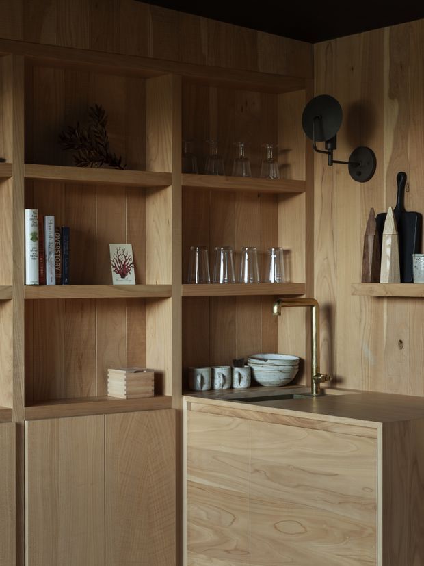 A warm, dimly lit built-in timber joinery wall with open display shelving, books, glassware, ceramics, concealed lower cabinets, and a compact integrated bar or kitchenette. A brushed brass wall-mounted tap and round black wall sconce provide contrasting accents. The cabinetry and shelving are made from pale-to-medium natural wood with visible grain, creating a refined, tactile interior.