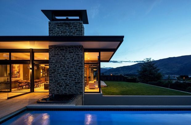 Twilight exterior view of a modern single-storey residence with extensive floor-to-ceiling glazing, a broad flat roof, and a prominent dark stacked-stone chimney rising through the roof. Warm interior lighting reveals dining and living areas, while a reflecting swimming pool occupies the foreground and a mountainous landscape forms the background.