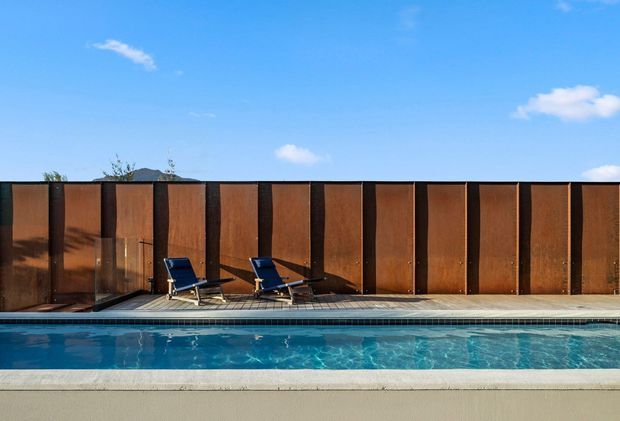 A sunny outdoor swimming pool beside a tiled pool deck, with two lounge chairs and a tall rust-brown timber or metal privacy screen forming the backdrop. The scene has a clean, modern architectural character, with blue sky and distant hills visible above the screen.