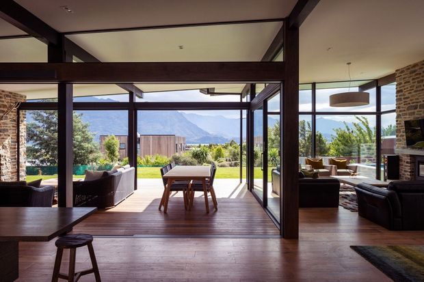 A spacious, open-plan interior with dark timber structural framing, exposed brick walls, extensive floor-to-ceiling glazing, and timber flooring. The central dining area opens directly to a landscaped outdoor space, while a living area with sofas and a fireplace is visible to the right. Mountain scenery is visible beyond the glazing. The design combines rustic natural materials with contemporary indoor-outdoor living.