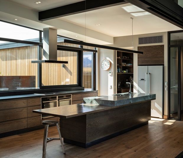 Contemporary kitchen with a large dark timber island, integrated seating, dark stone benchtop, extensive black-framed glazing, timber cabinetry, and a stainless-steel refrigerator. A high ceiling with exposed dark structural beams and pendant lighting creates a refined, light-filled space.