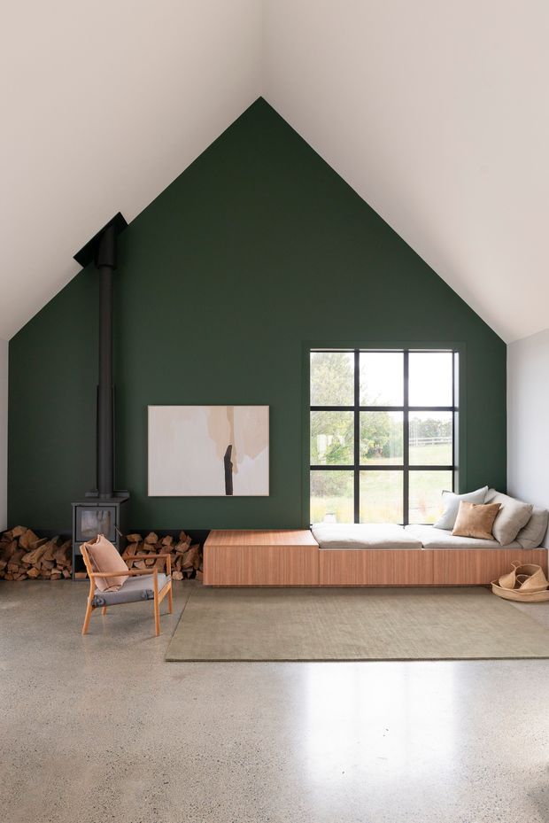 A refined living room with a dramatic dark forest-green gable wall rising to a pitched ceiling. The room features a black wood-burning stove with stacked firewood, a large black-grid window, a built-in timber window seat and cabinetry, neutral upholstered cushions, a timber-framed lounge chair, abstract wall art, and a muted rug over a polished concrete floor. The interior is warm, minimalist, and natural, with strong vertical and geometric architectural lines.