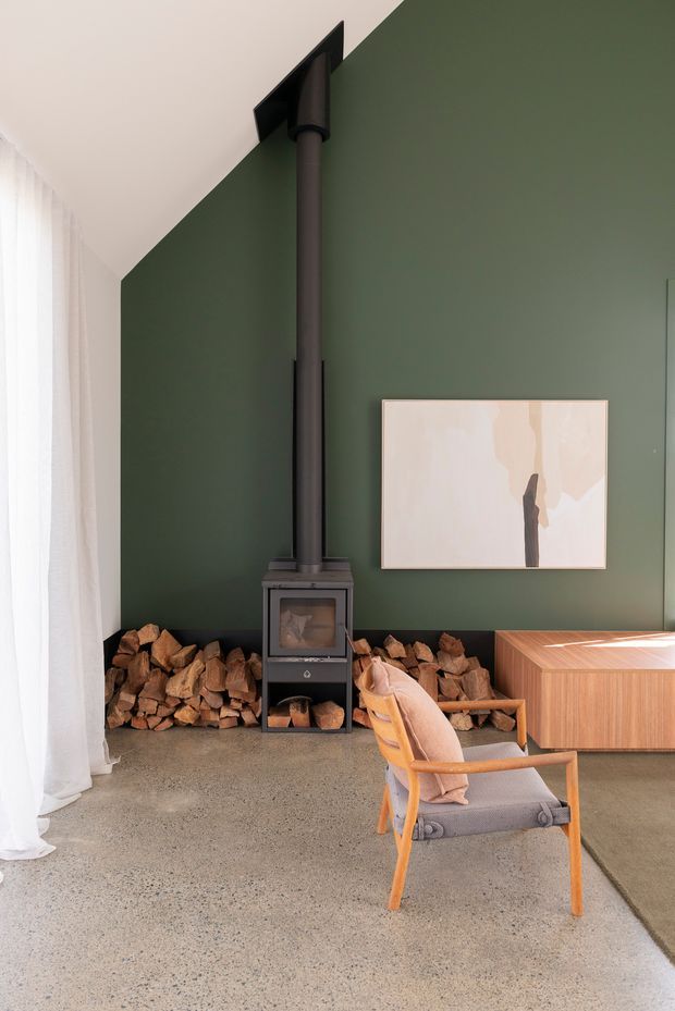 A refined living room with a dark green feature wall, freestanding black wood-burning stove and flue, stacked firewood, sheer white curtains, polished concrete flooring, and a warm timber credenza. A mid-century-inspired lounge chair and neutral artwork complete the calm, contemporary interior.