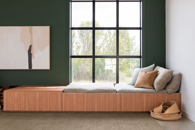 A calm, contemporary living room featuring a long built-in timber window seat beneath a large black-framed gridded window. The seat has pale cushions and neutral pillows, with a woven basket on the floor and abstract artwork on a deep green feature wall. The room has a soft neutral carpet, white side walls, and abundant natural light.