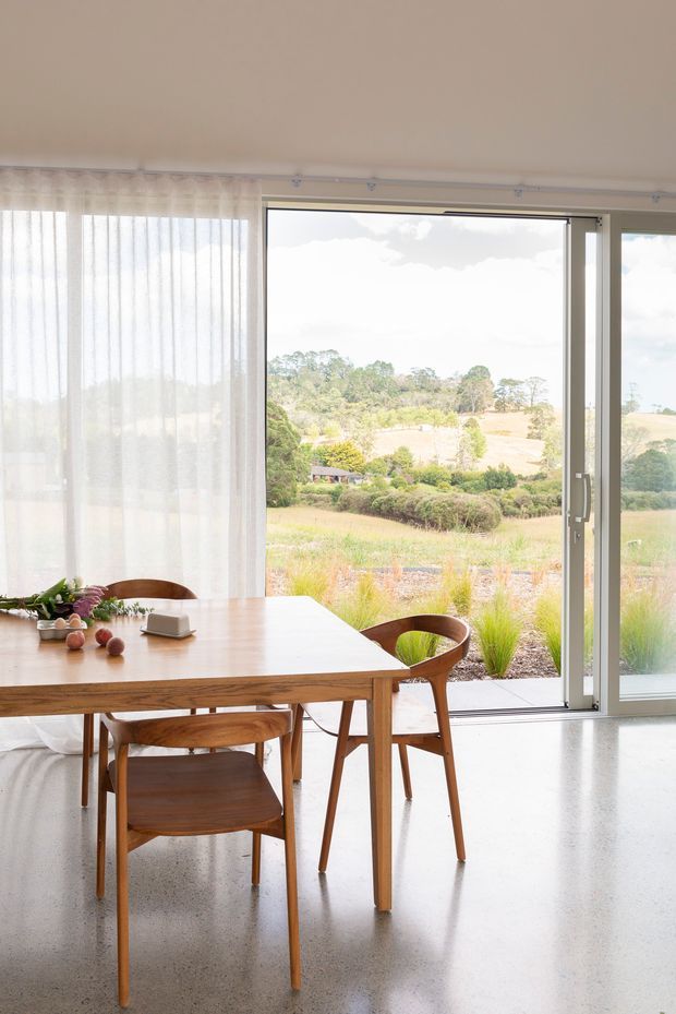 Bright dining area with a simple timber dining table and matching mid-century style wooden chairs, positioned beside full-height glazed sliding doors. Sheer curtains soften the daylight, while a rural landscaped setting is visible outside. The polished concrete floor and restrained neutral interior create an airy, natural atmosphere.