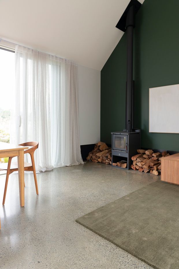 Bright, refined living area with a dark forest-green feature wall, freestanding black wood-burning stove and flue, stacked firewood, pale sheer curtains over large glazing, timber dining furniture, a low timber storage cabinet, and a muted rectangular rug over a polished grey concrete or terrazzo floor.