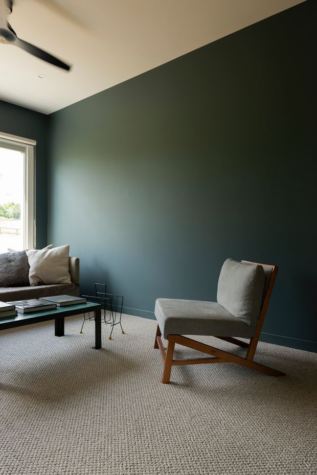 A refined living room with deep blue-green painted walls, a light ceiling, broad textured carpet, and understated mid-century-inspired furniture. A low dark coffee table, neutral sofa, sculptural lounge chair, and minimalist ceiling fan create a calm, restrained composition. Daylight enters through the tall window at left.