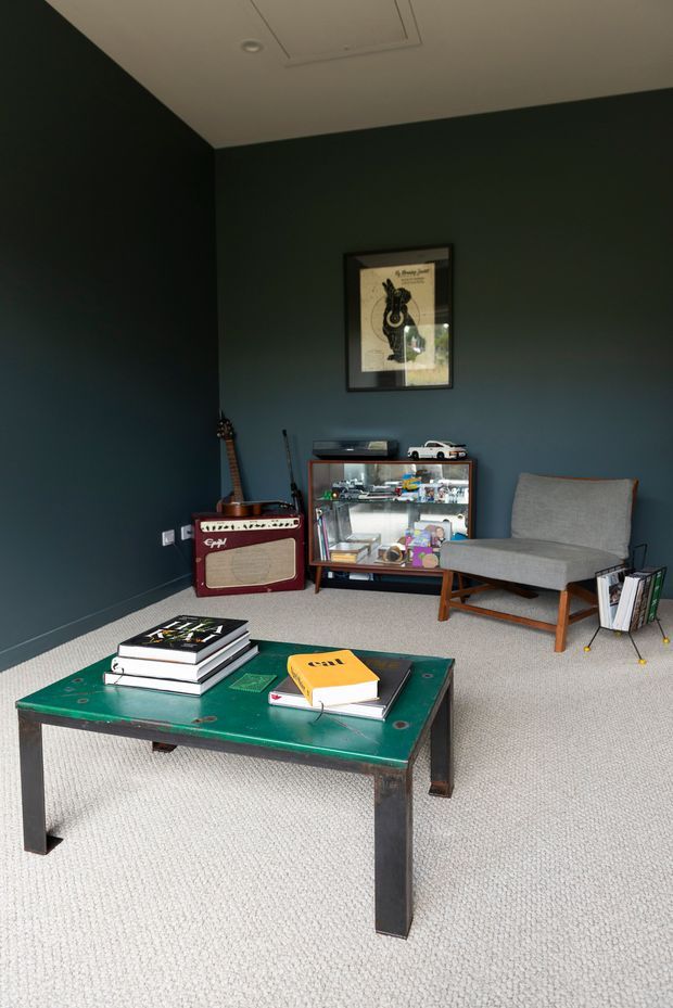 A moody, compact music- and hobby-themed sitting room with deep charcoal-blue walls, a light carpeted floor, and a low distressed green metal coffee table covered with books. A grey upholstered timber-framed lounge chair sits beside a glass-front display cabinet filled with collectibles, with a red guitar amplifier and guitars along the back wall. Framed artwork and small decorative objects complete the space.