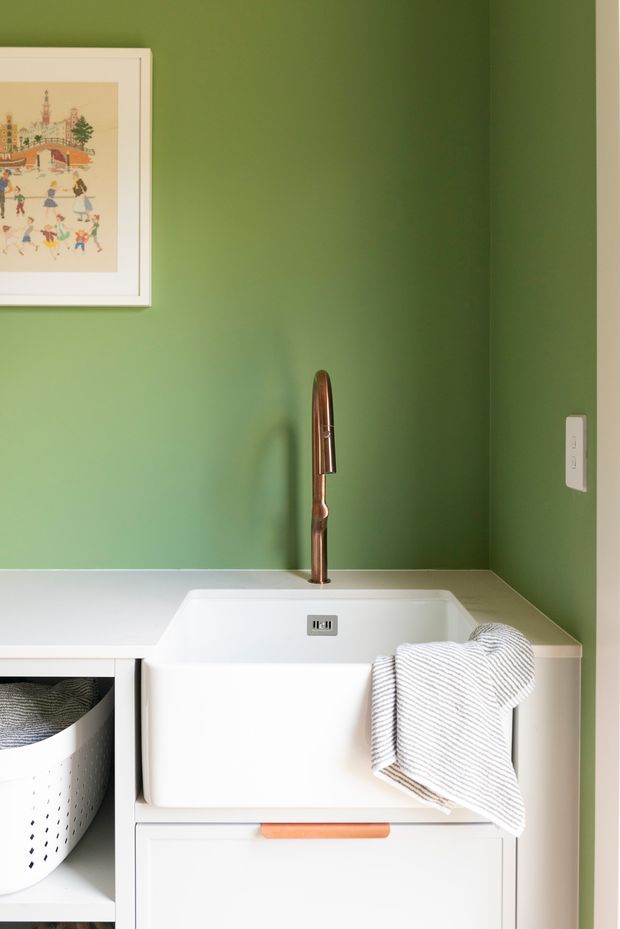 Close-up of a compact laundry area with a white farmhouse-style utility sink set into pale cabinetry, a brushed copper gooseneck tap, and a striped hand towel. The walls are painted a rich green, with a framed artwork and a white electrical outlet visible. A laundry basket is partly visible below the adjacent benchtop.