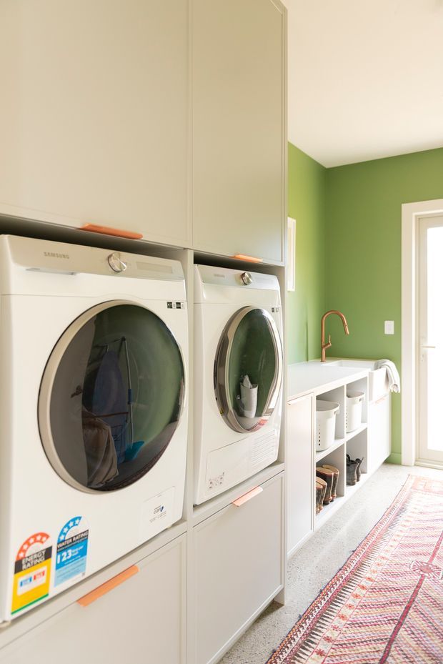 Bright, compact contemporary laundry room with stacked white front-loading washer and dryer integrated into tall pale cabinetry. A white benchtop with open storage cubbies runs along the green-painted wall, featuring a copper-toned gooseneck tap and utility baskets. A glazed door provides strong natural light, while a patterned runner adds warmth to the practical space.