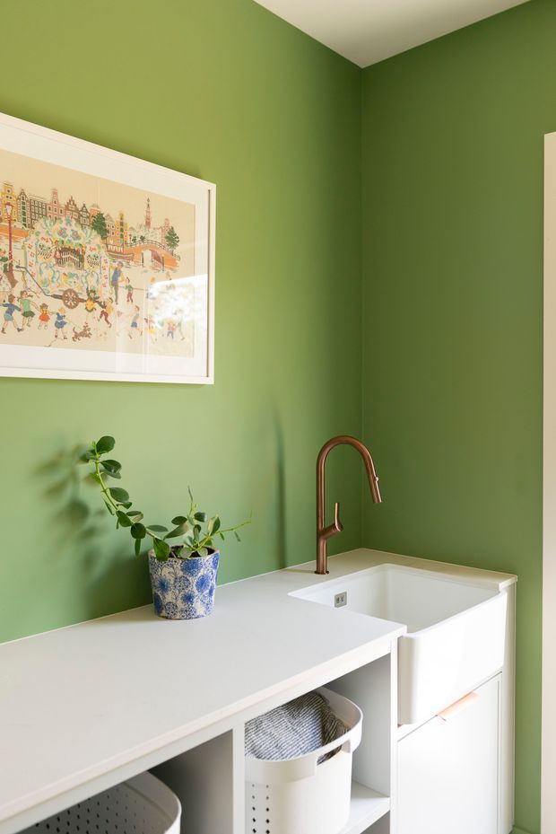 Compact laundry room with vivid green painted walls, white built-in cabinetry and benchtop, a deep utility sink with a copper gooseneck tap, open storage containing laundry baskets, a potted plant, and framed artwork.