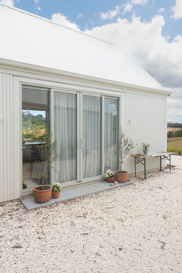Exterior view of a small white corrugated-metal building with a steep pitched roof, full-height glazed sliding doors, and a simple gravel forecourt. Potted plants and a narrow outdoor table sit beside the entrance, with open rural fields visible beyond.