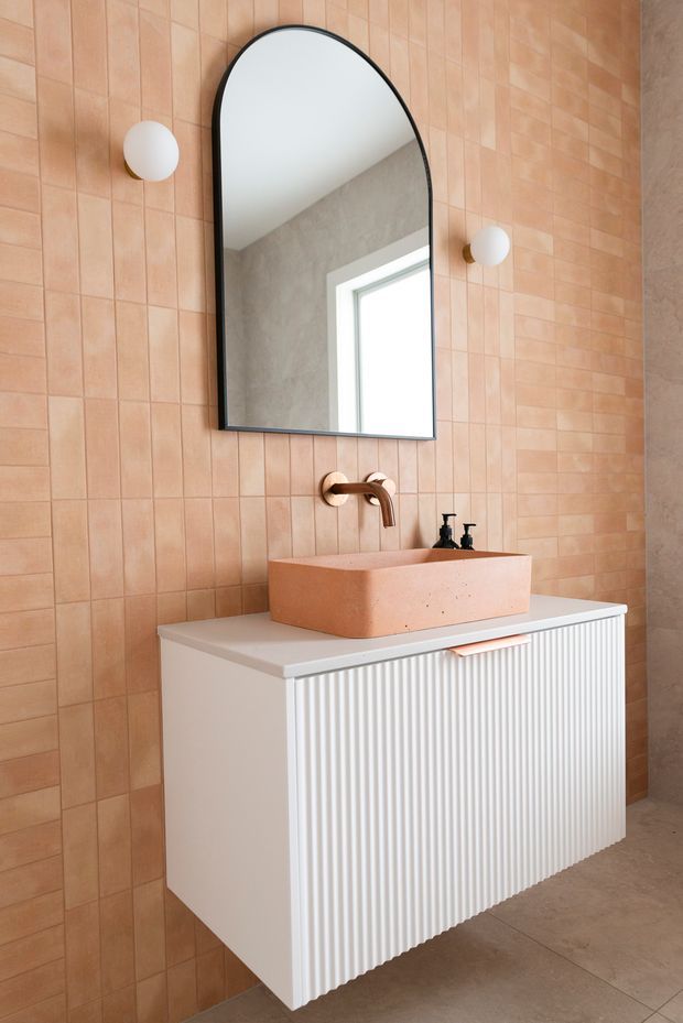 A contemporary bathroom vanity with a fluted white floating cabinet, pale terrazzo-like countertop, and a peach-toned rectangular vessel basin. The wall is clad in warm peach ceramic tiles, complemented by a bronze wall-mounted faucet, arched black-framed mirror, and spherical wall sconces. A concrete-toned floor and adjacent textured wall complete the warm, refined scheme.