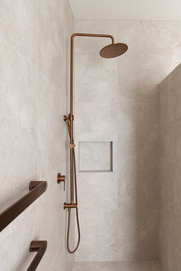 Close-up view of a contemporary walk-in shower clad in large-format pale beige stone-look tiles. The shower features a bronze/copper-toned exposed shower rail with a round overhead showerhead, handheld hose, recessed wall niche, and matching wall-mounted grab rails. The space is minimal, warm, and refined.