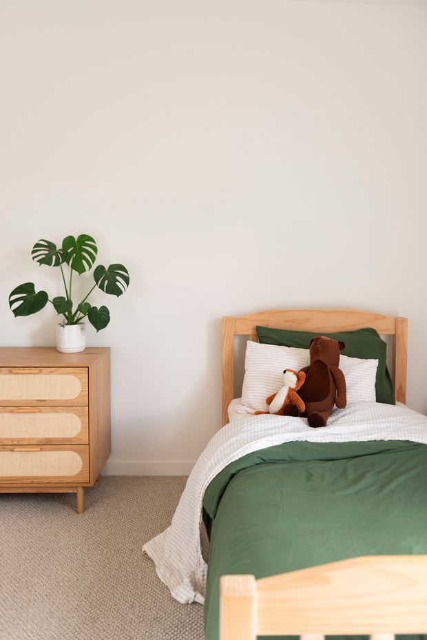 A warm, softly styled children's bedroom featuring a light timber bed with green bedding, a plush monkey toy, a matching timber dresser, a potted monstera plant, neutral walls, and carpeted flooring.