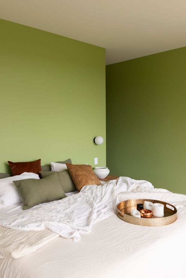 A cozy bedroom with a low bed dressed in rumpled white textured bedding and layered olive green, rust, and neutral cushions. The walls are painted in contrasting shades of muted olive green, with a white ceiling, simple wall light, and bedside vessel. A round hammered-metal tray with candles sits on the bed. The room has a calm, earthy and contemporary feel.