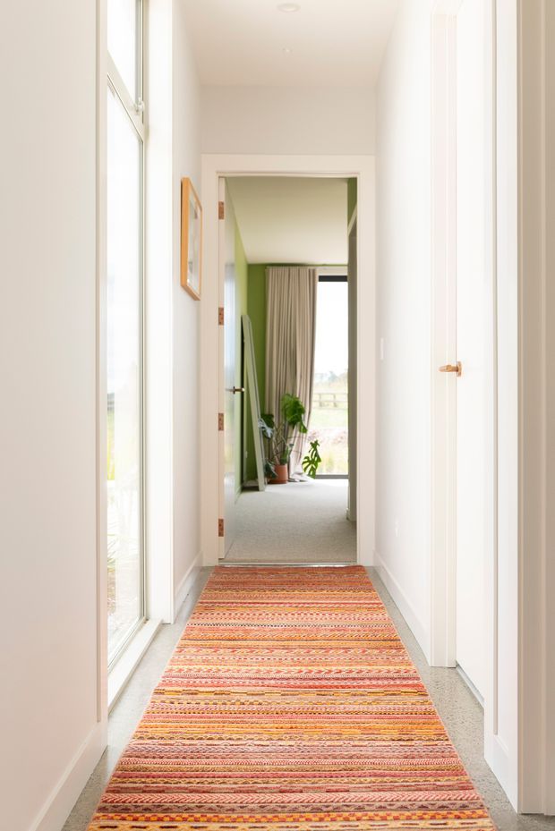 A bright, narrow residential hallway viewed toward an adjoining room. The corridor has white walls and doors, tall glazed openings along the left side, pale carpeted flooring, and a long multicoloured striped runner. A green-accented room with curtains, plants, and a large window is visible at the end.