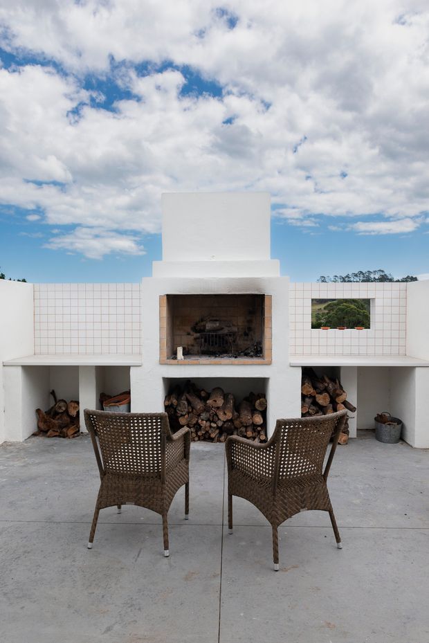 A minimalist outdoor courtyard or terrace featuring a large white masonry fireplace with a chimney, integrated open firewood storage, and white square-grid tiled walls. Two woven brown lounge chairs face the fireplace on a broad concrete floor. A small rectangular opening provides a view of landscaped greenery, while a bright blue sky with clouds is visible above the surrounding walls.
