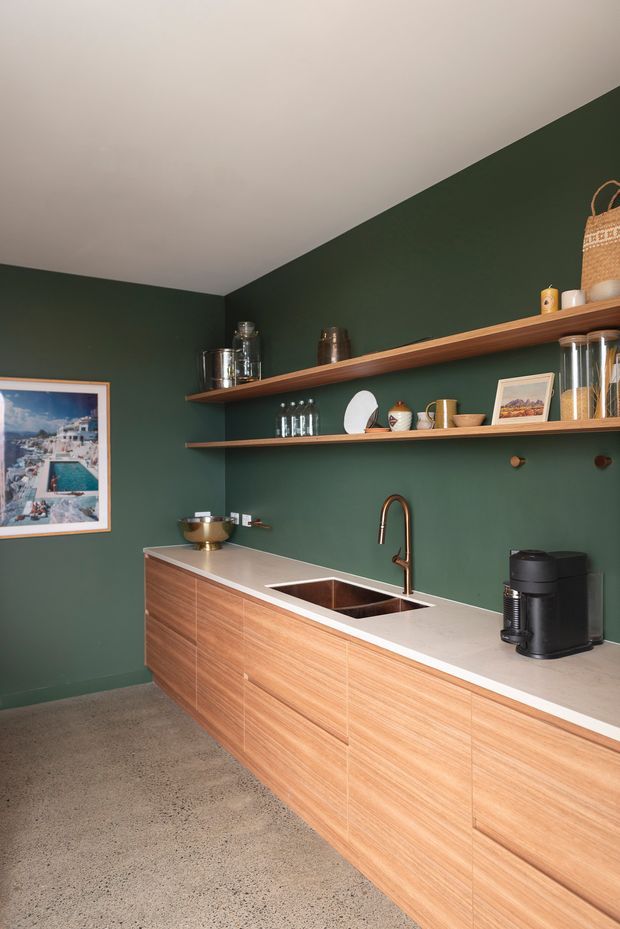 A compact contemporary kitchenette with deep forest-green walls, a long timber-grain built-in cabinet run, pale stone benchtop, integrated sink, and copper-toned gooseneck tap. Two open timber shelves display glassware, ceramics, framed artwork, and small decorative objects. The space has a polished concrete floor, a large framed coastal image, and warm natural styling with a clean, linear composition.