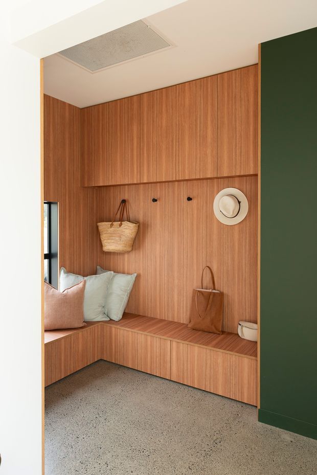 A compact built-in entryway or hallway nook featuring full-height warm timber wall panelling, integrated L-shaped bench seating with storage below, a narrow shelf, and wall hooks for hats and bags. The space includes soft cushions, woven and leather bags, a dark green adjacent wall, a white ceiling with a recessed air vent, and a speckled polished concrete floor. A slim window provides natural light at the left.