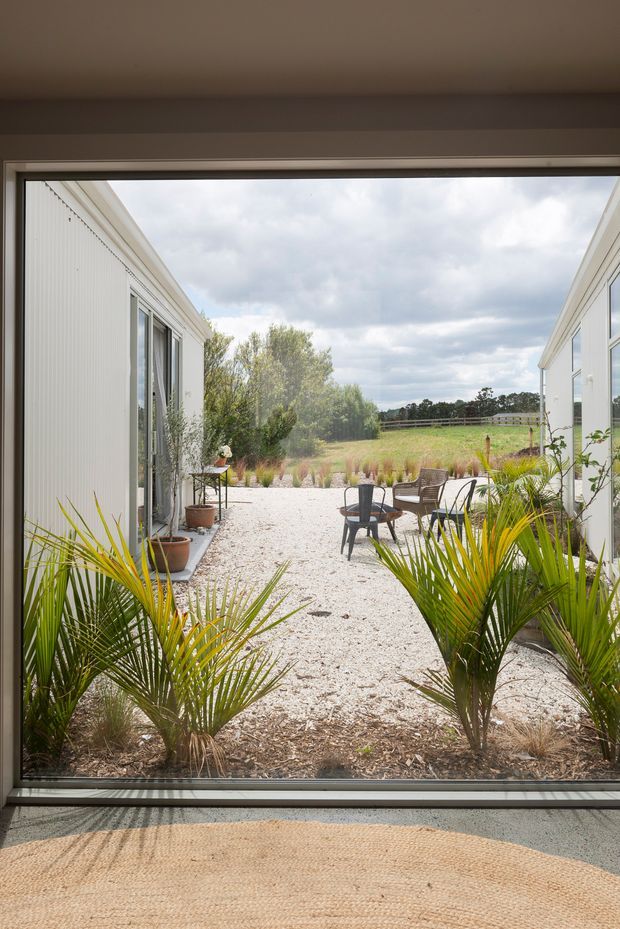 View from an interior through a large floor-to-ceiling glazed opening toward a gravel courtyard between two white-clad building wings. The courtyard contains potted plants, palms, outdoor seating and a small table, opening onto a grassy rural landscape beneath a cloudy sky. A woven rug and interior threshold are visible in the foreground.