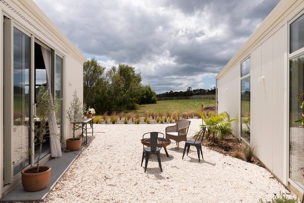 A bright landscaped courtyard between two single-storey white-clad building wings. Full-height glazed doors and windows open onto a pale gravel terrace furnished with a small round table, chairs, and potted plants. Low ornamental planting borders the terrace, with open rural fields and a dramatic cloudy sky beyond.