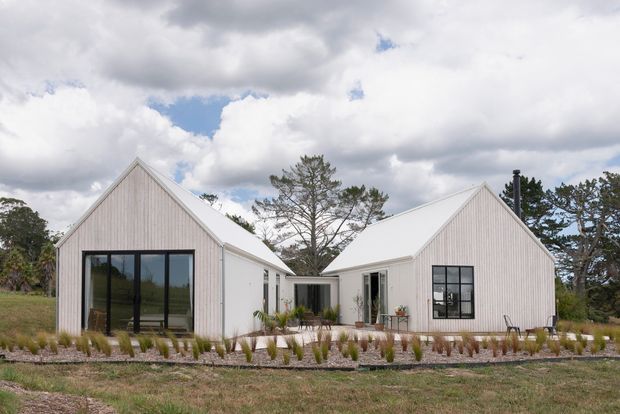 Exterior view of a contemporary single-storey residential complex composed of two white-clad gabled wings linked by a recessed central entry or courtyard. The buildings feature steep pitched roofs, vertical light-coloured cladding, dark-framed floor-to-ceiling glazing, and a landscaped gravel forecourt with ornamental grasses and outdoor seating. The rural setting and mature trees are visible around the home.