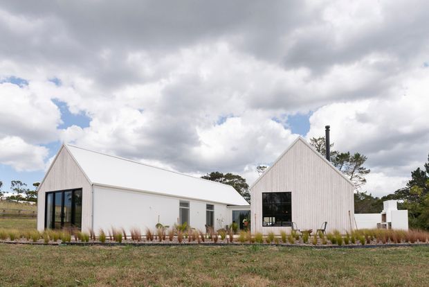 A minimalist white single-storey rural house composed of two steeply gabled volumes, with vertical light-coloured cladding, crisp white metal roofing, large dark-framed windows, and a landscaped garden foreground. The home sits in an open rural setting beneath a dramatic cloudy sky.