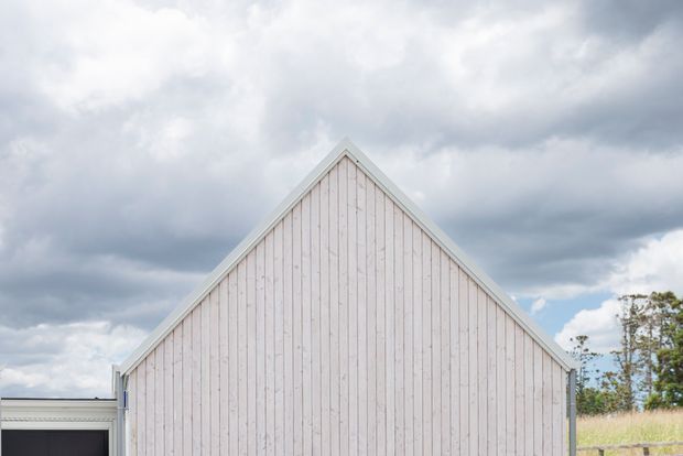 Exterior architectural view of a simple tall gabled structure with pale vertical timber cladding, white trim, and a dark garage or opening partially visible at the left. The building is set against a cloudy sky with sparse vegetation in the background.