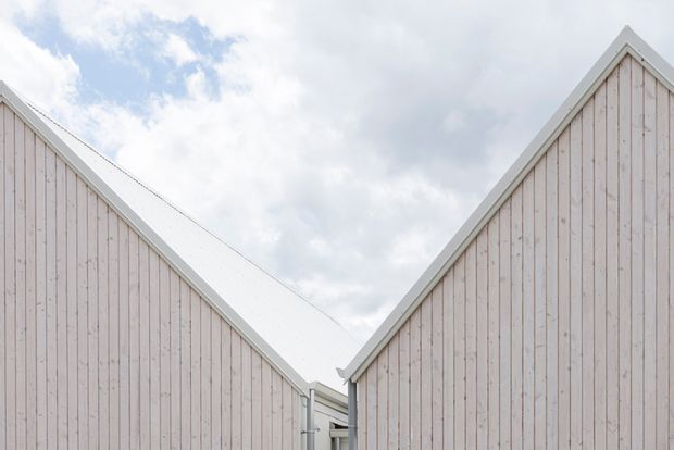 Exterior architectural detail showing two steeply pitched, gabled volumes clad in pale vertical timber boards with crisp white roof edging and trim. A narrow connection or passage is visible between the buildings, set against a cloudy sky.
