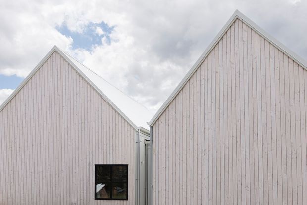 Exterior architectural view of two adjacent steeply gabled forms clad in pale vertical timber boards. A small dark-framed window is visible on the left volume, with a narrow glazed or metal-lined connection between the buildings. The scene is photographed under a partly cloudy sky, emphasizing the simple repetitive gable composition and light facade.