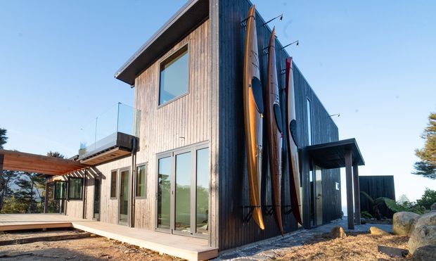 Exterior view of a contemporary multi-level home with dark vertical timber cladding, extensive glazing, a projecting entrance canopy, and a timber deck. Several surfboards are displayed vertically against the side facade, with landscaped rocky ground and a clear sky surrounding the building.