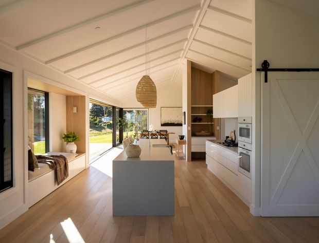 Bright open-plan kitchen and dining area with a central white island, integrated white cabinetry, timber flooring, large glazed openings, and a pitched ceiling with exposed painted rafters. A woven pendant light hangs above the island, while a white barn-style sliding door and built-in window seat add character. The space opens directly toward a landscaped exterior.