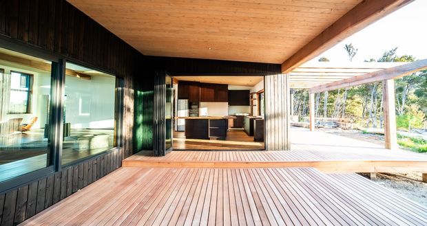 A modern timber-clad home opening onto a broad timber deck and covered outdoor living area. The image looks across the deck toward a compact kitchen, with extensive glazing on the left and a strong indoor-outdoor connection. Dark vertical timber wall cladding contrasts with warm timber ceilings, beams and decking, while a pergola frames the landscaped exterior.