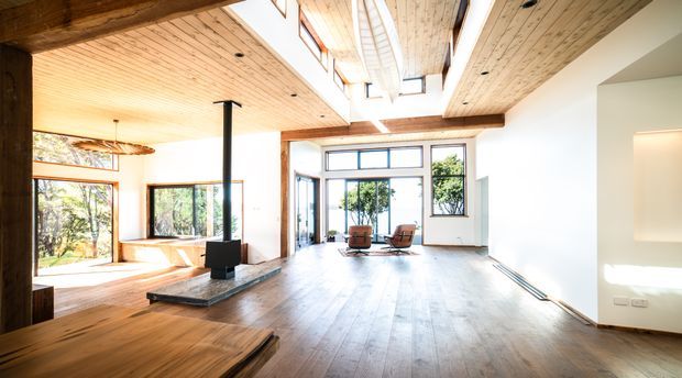 Spacious, light-filled open-plan living area with extensive timber flooring and timber-lined ceilings, exposed heavy timber beams, clerestory glazing, and large windows overlooking greenery. A central black wood-burning stove sits on a raised stone hearth, with a small seating area near the rear glazing. The room has a warm contemporary-rustic character and a strong connection to the outdoors.