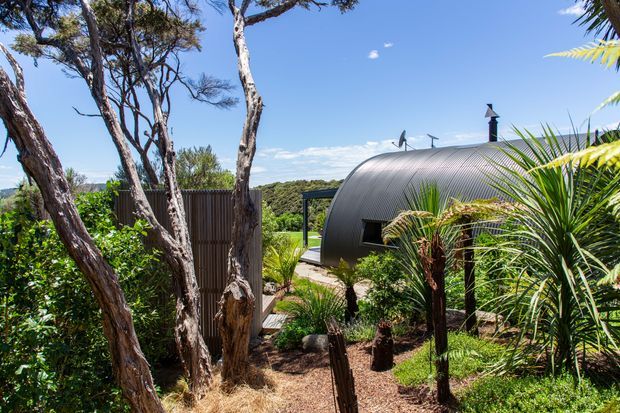 Exterior view of a compact curved-roof structure set within a landscaped garden. The building has dark corrugated metal cladding, a rounded barrel-like roof form, small dark-framed windows, and a chimney or vent. Native-looking trees, palms, rocks, and dense planting surround a gravel or mulch pathway, with a blue sky and distant vegetation beyond.
