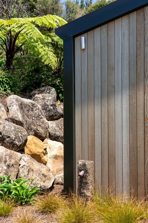 Close-up exterior view of a compact structure set within a lush, rocky landscape. The facade features vertical weathered timber boards framed by bold black metal trim, with a sloping roof edge and a small wall light. Native ferns, rocks, grasses, and a stone water spout or fixture reinforce the natural garden setting.