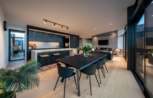 A contemporary open-plan kitchen, dining and living area with a long dark dining table and modern chairs in the foreground. The kitchen features a large island with dark vertical-panel cabinetry, a pale benchtop and integrated lighting. Dark built-in cabinetry and a wall-mounted television define the living area, while full-height black-framed glazing lines the right side. Light timber flooring, neutral walls, recessed lighting and indoor greenery create a refined, warm modern interior.