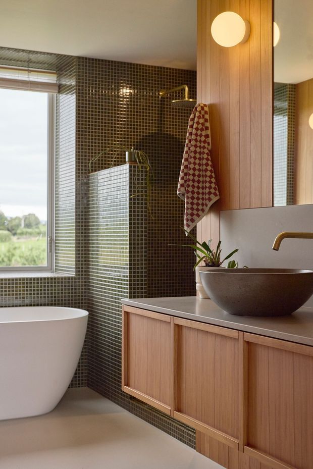 A warm, contemporary bathroom with a freestanding white bathtub beside a large window, dark small-format tiled walls, and a timber vanity with a stone benchtop. A round vessel basin, brass tapware, globe wall lights, a timber wall panel, and a patterned towel add refined natural detail.