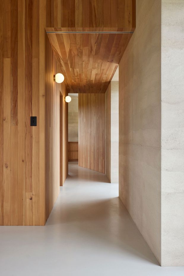 A minimalist interior hallway with extensive warm timber wall and ceiling lining, pale smooth concrete or plaster walls, and a light polished floor. The corridor is articulated by recessed openings and a timber-clad projecting volume, with small spherical wall lights providing warm illumination.