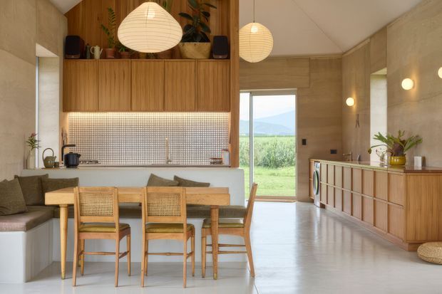 A warm, open-plan kitchen and dining area with a built-in timber dining banquette, a rectangular timber dining table, woven-seat dining chairs, and a compact kitchen island. The room features extensive vertical timber cabinetry and wall panelling, a small light mosaic tile splashback, pale concrete-look walls and floor, pendant lighting, indoor plants, and a glazed opening to a lush landscape beyond. A timber-fronted built-in cabinet runs along the right wall beneath round wall lights.