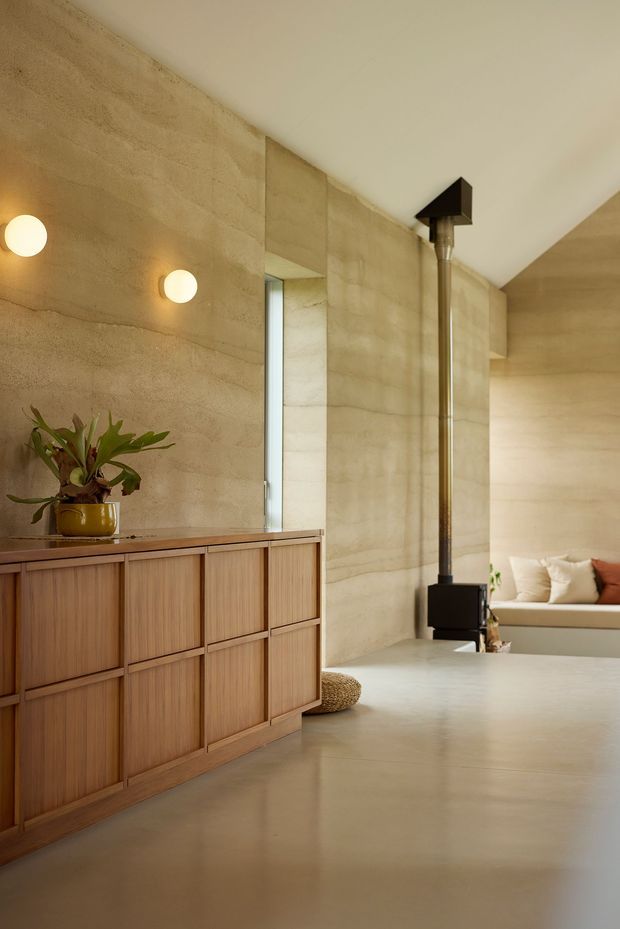 Warm, minimalist living room with monolithic beige concrete or rendered walls, a smooth polished concrete floor, and a low timber credenza with vertically grooved cabinet fronts. A freestanding black wood-burning stove with a tall flue is positioned near a recessed sitting area containing a cream sofa. Soft globe wall lights, a potted plant, and warm natural materials create a calm, refined interior.