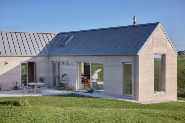 Exterior view of a contemporary single-storey gable-roof house with pale masonry walls, a dark standing-seam metal roof, large vertical windows and glazed doors, and a small outdoor dining patio opening onto a lawn.