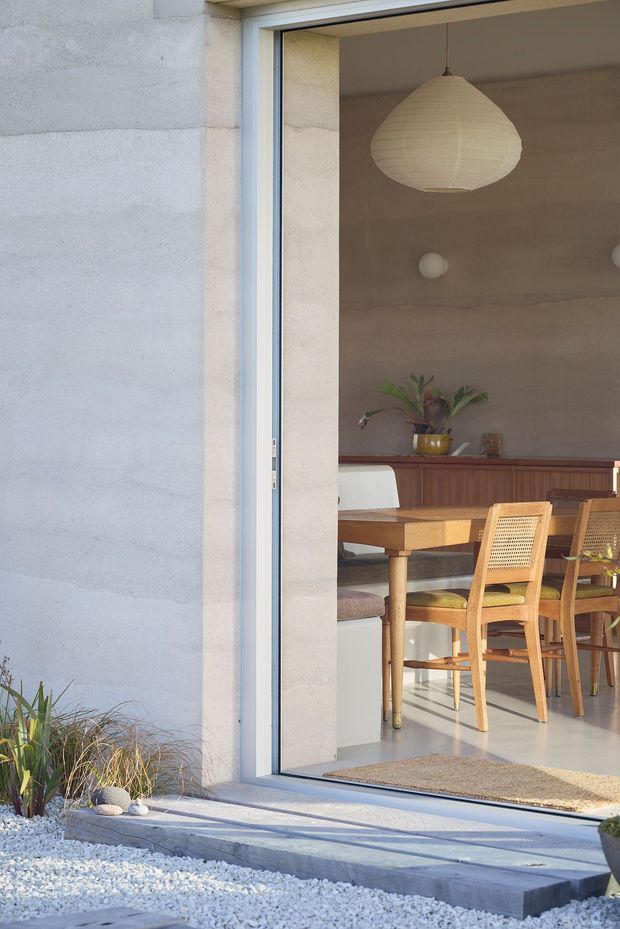 View from a pale rendered exterior through a large glazed sliding door into a warm, minimalist dining area. The room contains a timber dining table and woven timber dining chairs, a long timber sideboard, a sculptural paper pendant light, wall sconces, and restrained natural styling. White gravel landscaping and a shallow concrete threshold frame the exterior.