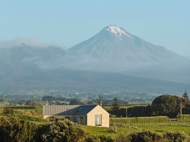 A small contemporary rural house with a simple gabled roof sits amid cultivated fields and hedges, with a large snow-capped volcanic mountain rising in the misty background. The scene has a calm agricultural landscape setting and clear views toward the mountain.