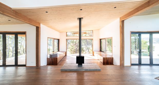 Bright open-plan living space with a timber-lined ceiling, exposed heavy timber beams and posts, warm timber flooring, and extensive glazed doors and windows overlooking a leafy outdoor setting. A central black freestanding wood-burning stove sits on a stone hearth, with built-in timber window seating along the sides.