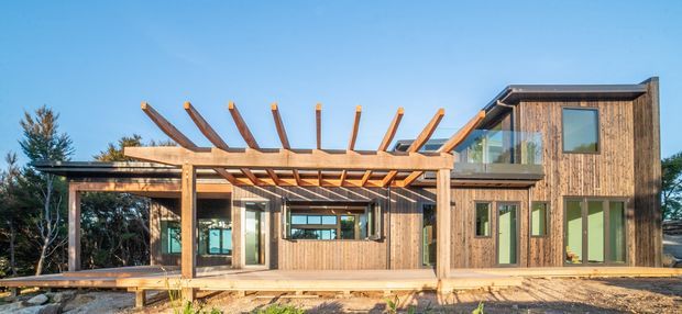 Exterior view of a contemporary two-storey timber-clad house with a large exposed timber pergola, broad glazing, and a low-slope roofline. The home sits in a landscaped setting beneath a clear blue sky.