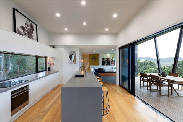 Bright open-plan contemporary kitchen with a long central island, integrated white cabinetry, timber flooring, and large black-framed glazing opening onto a timber deck. A dining area and living space are visible beyond the kitchen, with a leafy landscape outside.
